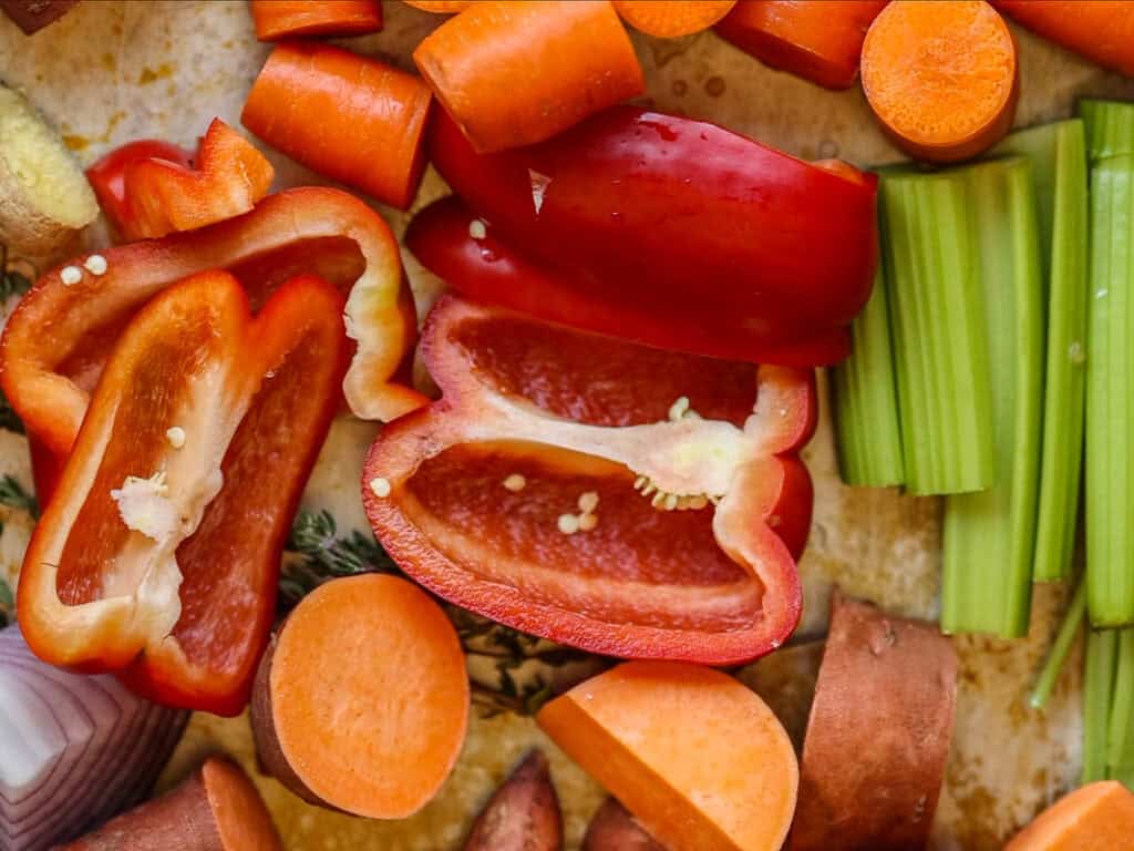 A colorful assortment of chopped vegetables, including red bell peppers, carrots, celery, and sweet potatoes, displayed on a wooden surface.